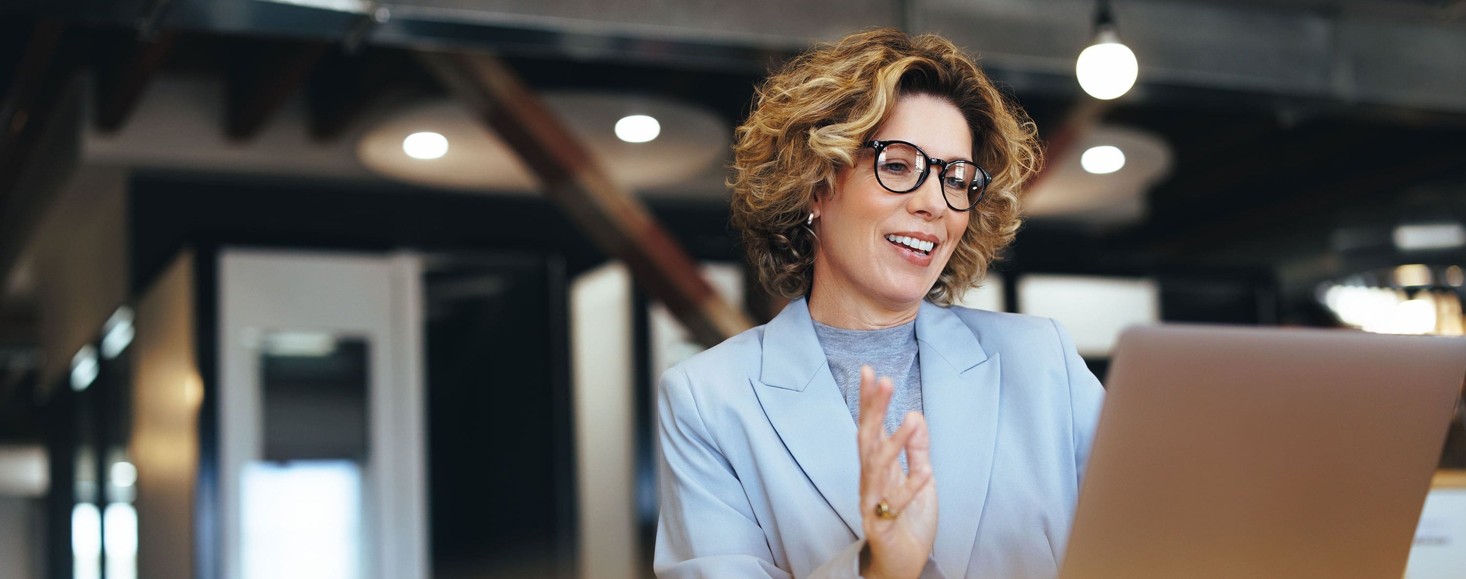 Frau mit Brille in hellblauem Blazer blickt im Büro auf einen Laptop.