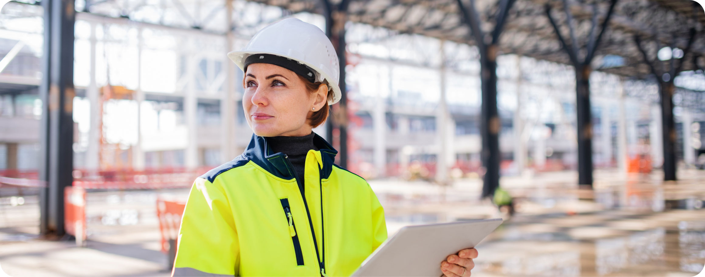  Eine Ingenieurin in Sicherheitskleidung und Helm steht in einer großen, im Bau befindlichen Halle und hält ein Tablet in der Hand.