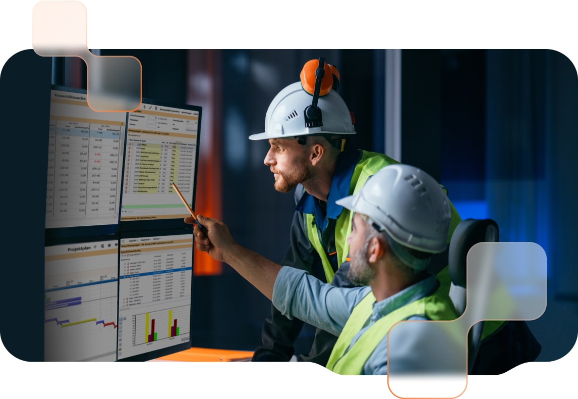 Zwei Bauingenieure mit Sicherheitshelmen und Warnwesten analysieren Daten auf mehreren Monitoren in einem Büro.