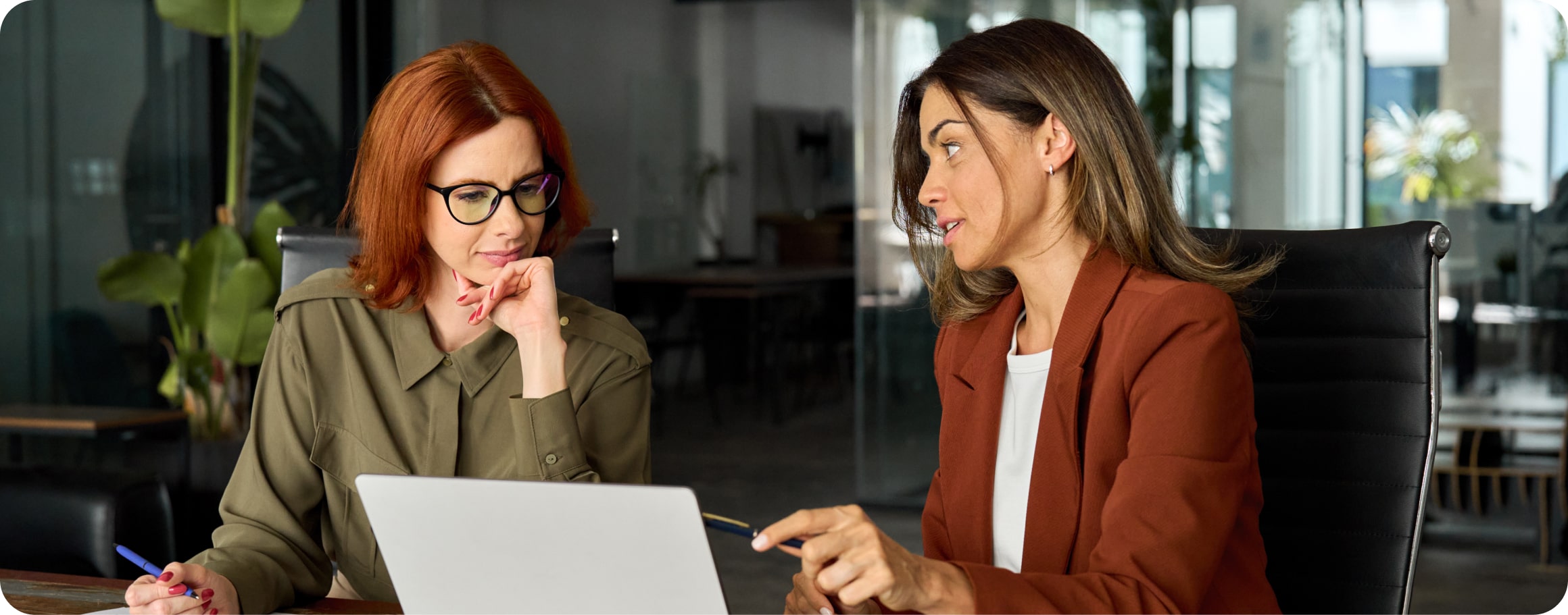 Zwei Frauen sitzen in einem Büro und unterhalten sich vor einem Laptop sitzend. 