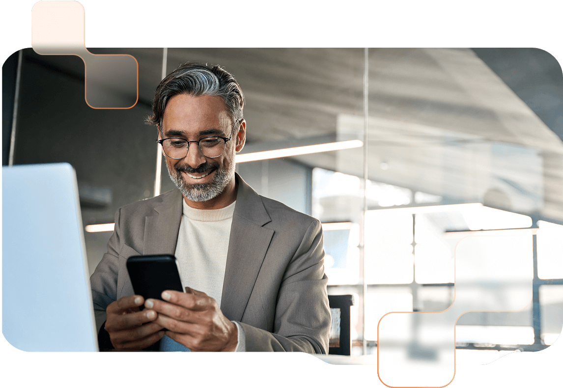 Mann mit Brille blickt lächelnd auf sein Smartphone, vor ihm steht ein Laptop.