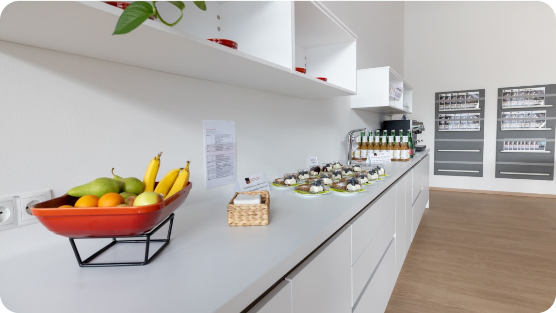 Moderner Akademiebereich mit einer weißen Theke, frischem Obst in einer Schale, kleinen Snacks auf Tellern und Getränken in Flaschen.