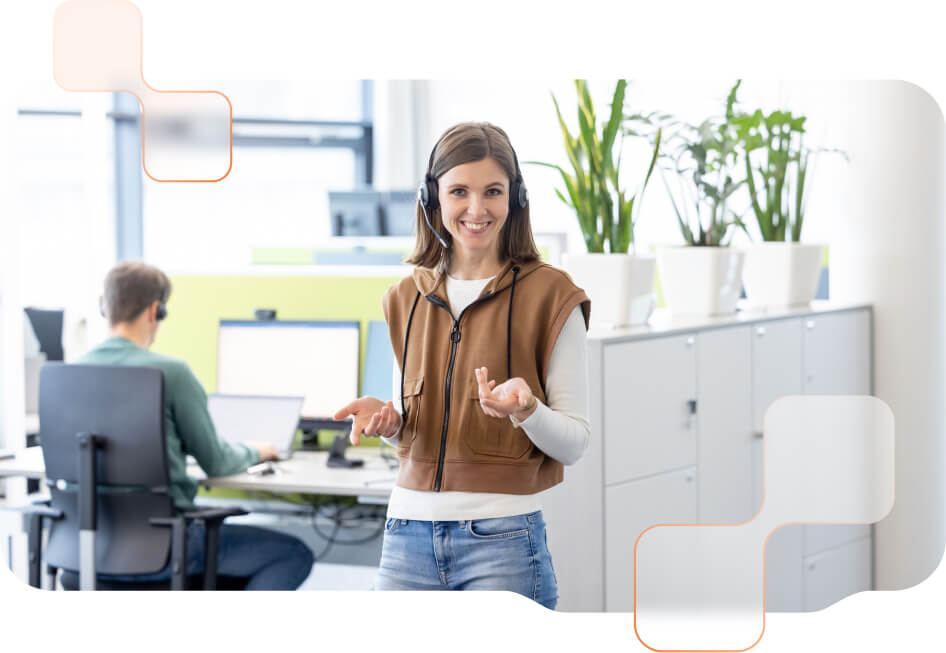 BMD Support Mitarbeiterin mit Headset lächelnd und stehend im Büro - im Hintergrund sitzt ein weiterer BMD Mitarbeiter beim PC.