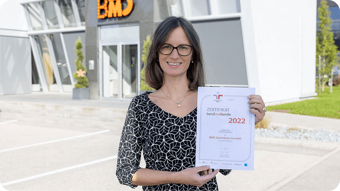 Eine BMD Mitarbeiterin steht vor dem BMD Gebäude und hält freudestrahlend ein Zertifikat in den Händen.
