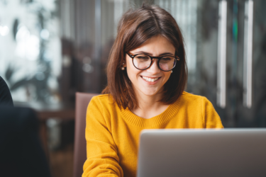 Eine Frau mit Brille und orangem Pullover sitzt vor aufgeklapptem Laptop. [© stock.adobe.com - leonidkos]