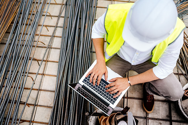 Das Bild zeigt einen Baufirmen-Mitarbeiter mit Helm, Warnweste und Laptop auf der Baustelle. [©stock.adobe.com - bogdanhoda]