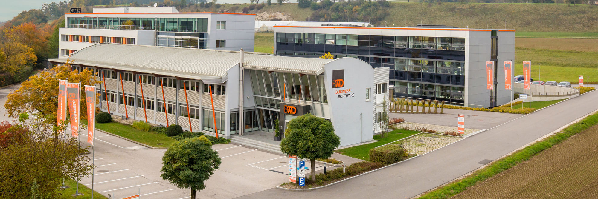 The company building of BMD Systemhaus GesmbH with large glass fronts, surrounded by parking lots and green landscape. Several orange flags with the company logo are displayed in front of the building.
