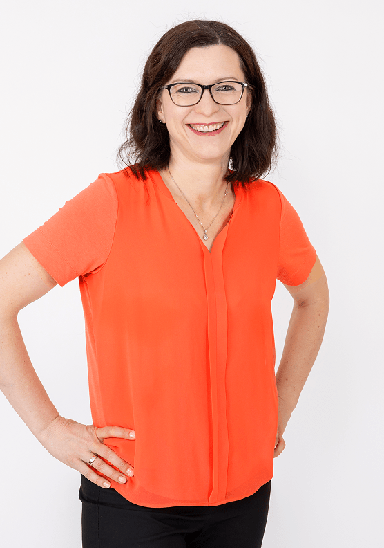 Eine Frau lächelt in die Kamera. Sie hat schwarze Haare und trägt eine orange Bluse und eine schwarze Hose.