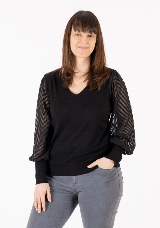 Eine Frau lächelt in die Kamera. Sie hat braunes, langes Haar mit Stirnfransen. Sie trägt eine schwarze Bluse und eine graue Jeans.