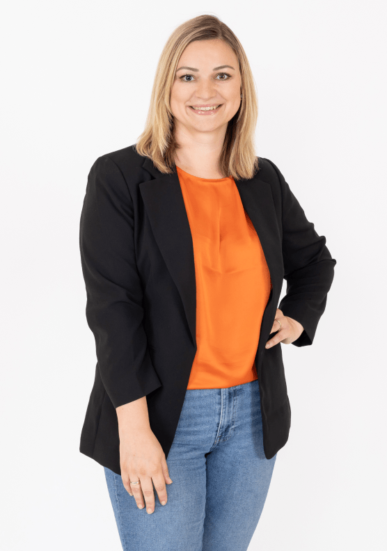 Eine Frau lächelt in die Kamera. Sie hat blondes, glattes Haar und trägt ein oranges Shirt mit schwarzem Blazer und einer Jeans.