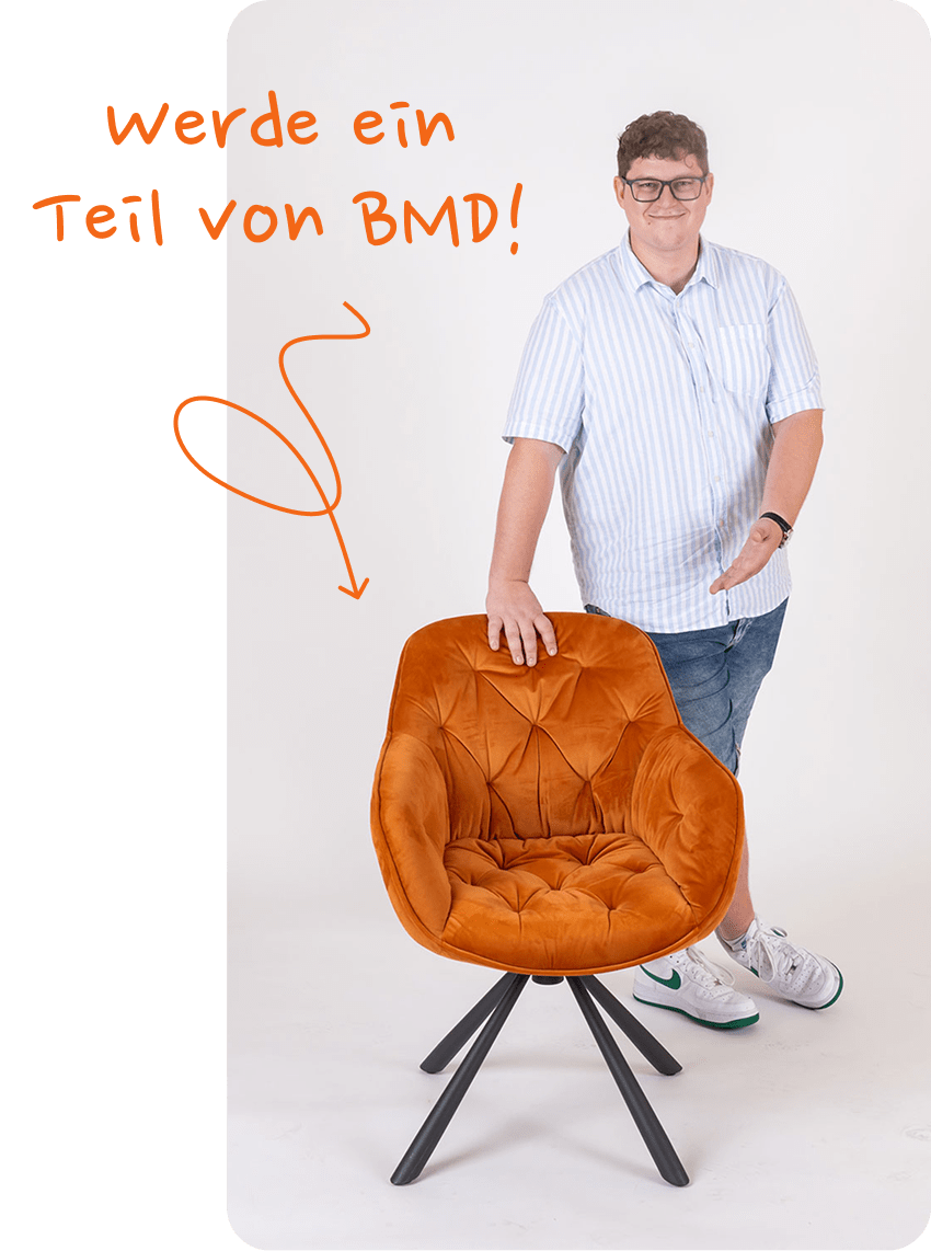 Ein junger Mann steht hinter einem orangen Sessel und zeigt auf die Sitzfläche