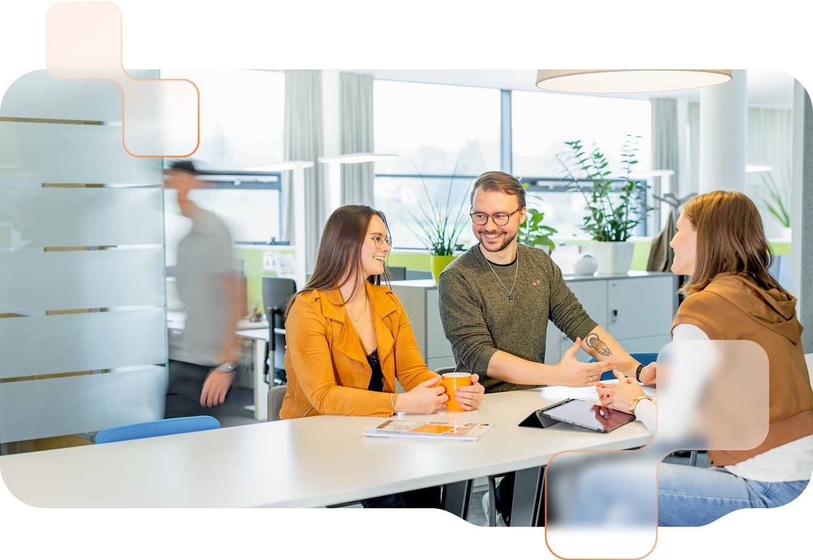 Drei Personen sitzen bei Besprechungstisch und unterhalten sich, während im Hintergrund jemand vorbeigeht. 